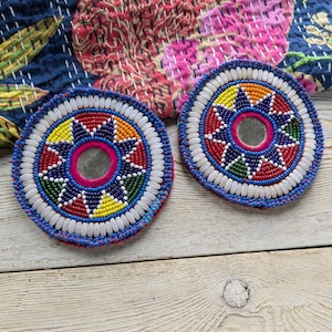 May include: Two round, beaded decorative pieces with a central mirror and colorful starburst patterns. The pieces feature white, red, blue, yellow, and green beads, with a blue border. The background includes a patterned textile.
