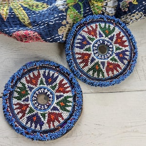 Peut inclure: Deux dessous de verre circulaires ornés de perles disposées en motif radial. Le design intègre des perles blanches, rouges, vertes et bleu foncé avec un petit miroir circulaire au centre de chaque pièce. Les bords sont finis avec des perles bleues.