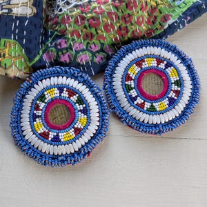 Puede incluir: Dos adornos circulares de cuentas con un anillo central rosa rodeado de patrones concéntricos de cuentas blancas, amarillas, verdes, rojas y azules. Los bordes exteriores están delineados con cuentas blancas y un borde de cuentas azules.