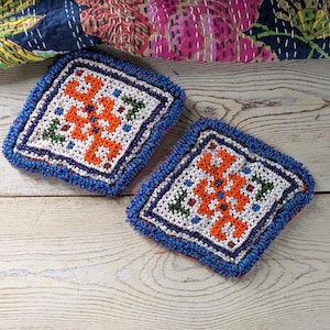 Peut inclure: Deux dessous de verre carrés perlés avec un motif géométrique. Les dessous de verre présentent un fond blanc avec un motif orange, vert et bleu. Les bords sont ornés de perles bleues. Ils sont posés sur une surface en bois clair.