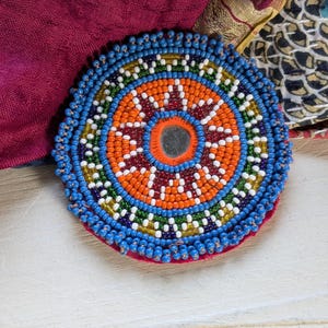 May include: A circular beaded patch featuring a central small mirror surrounded by concentric rings of orange, white, green, and blue glass beads. The outer edge is finished with a fringe of blue beads, creating a vibrant, multi-colored design.