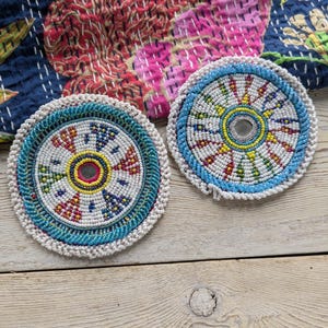 May include: Two round, beaded coasters with intricate designs. The coasters feature a central mirror surrounded by colorful beads in blue, yellow, red, and white. A turquoise border and a white beaded edge complete the design. The coasters are placed on a light wood surface.