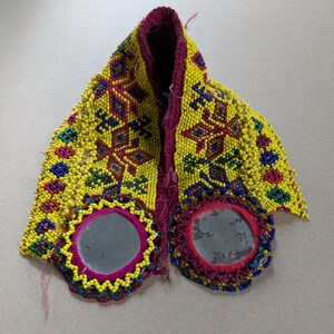 May include: A decorative textile accessory featuring a vibrant yellow beaded surface with intricate geometric patterns in blue, red, and green. The piece includes two circular mirrors framed by pink fabric and dense, multi-colored beadwork borders.