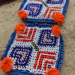 May include: Two square beaded items with intricate patterns in white, blue, orange, and red. Each item has a blue beaded border and four orange yarn pom-poms. The items are placed on a wooden surface.