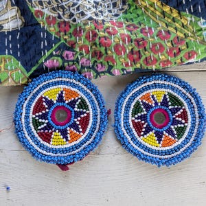 May include: Two circular coasters featuring intricate beadwork in blue, white, yellow, orange, green, and red. Each coaster has a small central mirror surrounded by a star-shaped pattern of beads, finished with a thick blue beaded border.