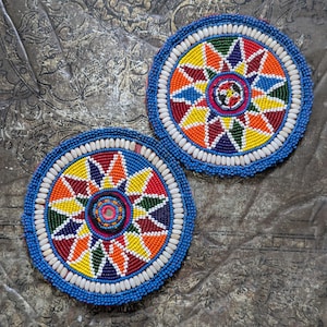 Peut inclure: Deux dessous de verre ronds et perlés avec un motif géométrique vibrant. Les dessous de verre présentent une bordure bleue, des perles blanches et un motif central de triangles et de formes colorées en rouge, jaune, orange et vert. Sur une surface métallique texturée.