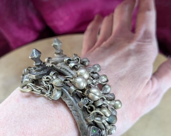 May include: A metallic cuff bracelet featuring a weathered silver-tone finish, adorned with multiple dangling spherical bells and two prominent pointed finials. The band displays etched geometric patterns and a small inset green stone.
