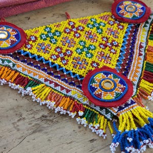 May include: A colorful, beaded textile with a triangular shape. The central design is yellow with a floral pattern, bordered by intricate beadwork in various colors. Red, circular accents with mirrors are placed at the corners. The edges are finished with beaded fringe.