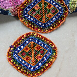Peut inclure: Deux dessous de verre perlés avec un motif géométrique. Les dessous de verre présentent une base rouge avec des perles orange, bleues, blanches et vertes. Le motif comprend un losange central et une bordure de petites perles blanches en forme de fleur.
