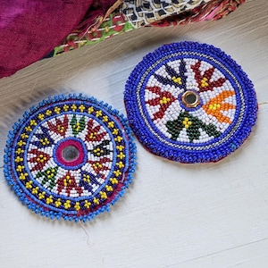 May include: Two circular beaded patches featuring intricate geometric patterns in blue, white, red, yellow, green, and orange glass beads. Each patch has a small reflective circular mirror at the center and a vibrant blue beaded border edge.