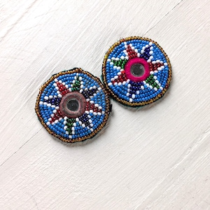 May include: Two circular patches featuring intricate beadwork in blue, white, red, and green. Each patch has a central circular mirror element. The beadwork is arranged in a starburst pattern radiating from the center, with a gold-toned beaded border.