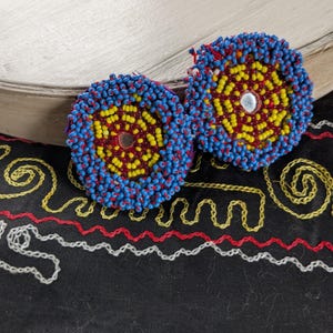 May include: Two circular earrings featuring intricate beadwork in vibrant blue, yellow, and deep red. Each earring has a small, reflective silver-toned center piece. The beads are arranged in concentric patterns, creating a textured, rounded surface.
