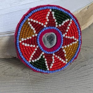 Puede incluir: Un parche circular de cuentas con un espejo central rodeado por un patrón de estrella de cuentas de vidrio rojas, amarillas y verdes. El diseño está delineado con cuentas azul claro sobre una base de tela rosa con costuras visibles.