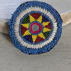 May include: A circular patch featuring a central mirror surrounded by concentric rings of beads. The design includes a star pattern in yellow, red, green, and blue, encircled by a ring of white oval beads and an outer border of small blue beads.