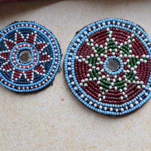May include: Two round, beaded decorative pieces. One is blue, red, and white, the other is red, green, and white. Both have a small mirror in the center. The beads are sewn onto a black backing.