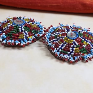 May include: Two colourful beaded round ornaments with a button in the centre. The ornaments are made of red, blue, green, yellow, and white beads.