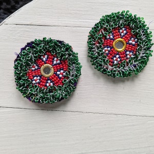 May include: Two round, beaded ornaments with a central mirror. The ornaments feature a red and white floral pattern, surrounded by green and silver beads. The background is a light-colored, textured surface.
