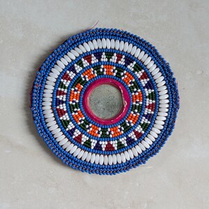 May include: A circular decorative wall hanging featuring a central mirror framed by concentric rings of multi-coloured glass beads. The design includes vibrant orange, green, purple, and red patterns, finished with an outer border of white beads.