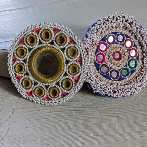 May include: Two circular embroidered patches featuring intricate beadwork and small mirrors. The left patch has a gold center ring surrounded by white beads and pink accents. The right patch features a blue center with multiple small mirrors and white beaded edges.