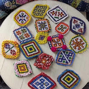 May include: A collection of colorful, beaded coasters in various geometric patterns. The coasters feature vibrant colors like red, blue, yellow, and green, with intricate designs. Some have pom-pom accents, showcasing a handcrafted aesthetic.