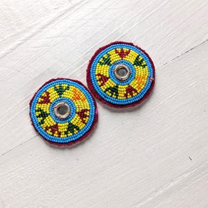 May include: Two circular patches featuring intricate beadwork in vibrant yellow, blue, green, and red patterns. Each patch has a small, reflective metallic center piece and is finished with a dark red fabric backing along the outer edge.