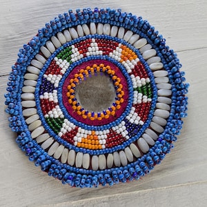 May include: A circular beaded medallion featuring a central mirror surrounded by concentric rings of orange, white, red, green, and blue glass beads. The outer edge is finished with a border of elongated white beads and small blue seed beads.