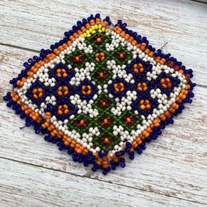 May include: A square, beaded textile with a geometric pattern. The design features a central green area with red and white floral motifs, surrounded by white, orange, and blue beads. The edges are outlined in blue beads.