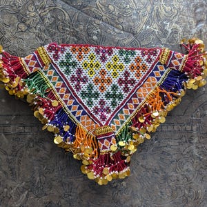 May include: A colorful beaded textile with a triangular shape, featuring a geometric pattern in white, green, orange, and yellow. The edges are adorned with gold-colored coins and beaded fringe. The background is a textured, dark surface.