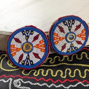 May include: Two circular beaded patches featuring a radial pattern of white, blue, orange, and dark red glass beads. Each patch has a small, reflective metallic center piece. The edges are finished with a dark red fabric border.