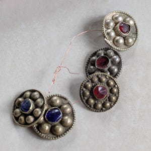May include: Four circular metal buttons connected by thin copper wire. Each button features a central faceted glass stone, two in deep red and two in dark blue, surrounded by a ring of small, raised metallic spheres with a tarnished finish.
