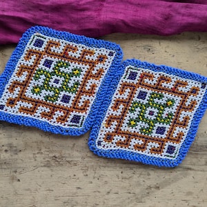 May include: Two square beaded coasters with a geometric pattern. The coasters have a blue beaded border and feature a central design in white, orange, green, and purple. They are placed on a wooden surface.