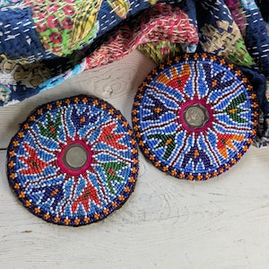 May include: Two circular coasters featuring intricate beadwork in a radial pattern of blue, red, green, and white glass beads. Each coaster has a central metallic coin-like insert surrounded by a bright pink border, set against a patterned fabric.