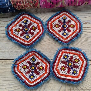 May include: Four beaded coasters with a geometric design. Each coaster features a central pattern of white, red, and yellow beads, framed by orange, red, and blue beaded borders. The coasters are arranged on a light wood surface.