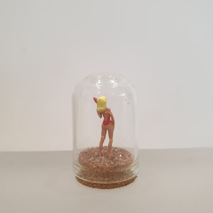 May include: A miniature figurine of a woman in a red swimsuit and yellow hat is inside a glass dome on a cork base. The dome is filled with sand.