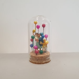 May include: A glass dome with a cork base contains a miniature family of three figures with colourful balloons. The figures are a woman holding a child, a man, and a child holding a balloon.