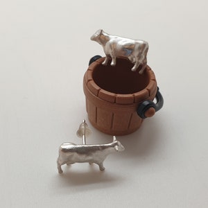 May include: A pair of silver cow earrings. One cow is standing on top of a small brown wooden bucket. The other cow is laying down.
