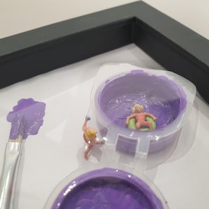 May include: Two miniature figures, one in a pool float and one holding a paintbrush, are placed in a purple paint container. The container is in a white frame with a black border.