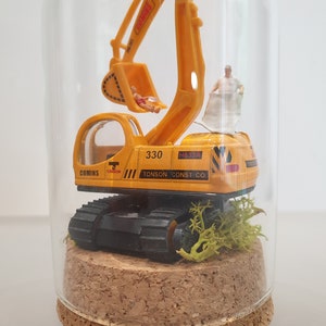 May include: A yellow toy excavator with black tracks and a clear cab is inside a glass dome. The excavator has the text "330", "TONSON CONST CO", and "COMINS" on the side. The dome is sitting on a cork base.