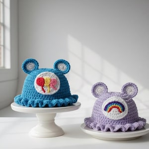 May include: Two handmade crochet hats. One is blue with a colorful patch and the other is lavender with a rainbow patch. Both hats have ear-like details and ruffled brims. They are displayed on white cake stands and plates.