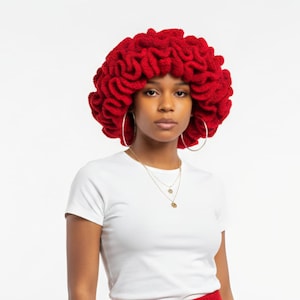 May include: A woman wearing a vibrant red, crocheted head covering. The headpiece has a textured, layered design. She is also wearing a white t-shirt, gold necklaces, and large hoop earrings. The background is plain white.