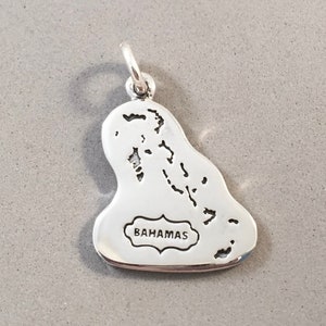 May include: A silver charm pendant in the shape of the Bahamas. The pendant features a detailed outline of the islands and the word "BAHAMAS" in a decorative frame. The charm has a loop for attaching to a necklace or bracelet.