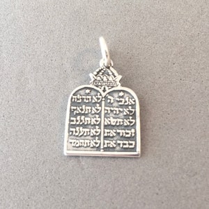 May include: Silver pendant featuring the Ten Commandments in Hebrew script. The pendant has a rectangular shape with rounded top edges and a crown design at the top. The pendant is suspended from a small loop.