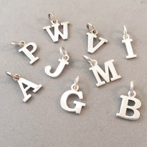 Könnte beinhalten: Eine Sammlung silberfarbener Buchstabenanhänger, jeweils mit einer kleinen Schlaufe zur Befestigung. Die Buchstaben umfassen A, B, G, I, J, M, P, V und W. Die Anhänger werden auf einer grauen Oberfläche präsentiert.