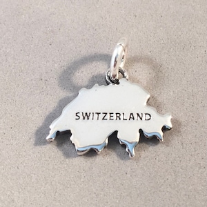 May include: A silver charm in the shape of Switzerland, with the country's name in black lettering. The charm has a loop at the top for attaching to a bracelet or necklace. The charm is on a gray background.
