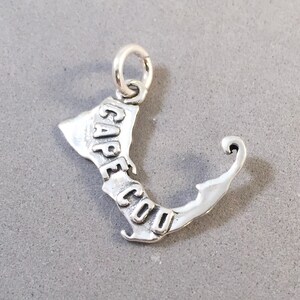 May include: A silver charm in the shape of Cape Cod, with the words "CAPE COD" engraved on the charm. The charm has a small loop at the top for attaching to a necklace or bracelet. The charm is on a gray surface.