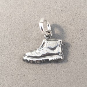 Può includere: Un ciondolo color argento a forma di scarpa da trekking. Lo stivale presenta trame dettagliate, tra cui lacci e una suola scanalata. Un piccolo anello è attaccato in alto per appenderlo. Il ciondolo è probabilmente in metallo.