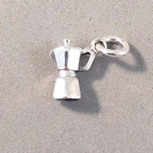 May include: A small, shiny silver charm in the shape of a stovetop espresso maker. The charm has a handle and a loop for attaching to a bracelet or necklace. The charm is highly detailed and reflects light.