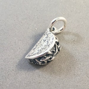 May include: A silver-colored taco charm with a textured surface. The charm has a detailed design, including the taco shell and filling. It features a small loop for attaching to a necklace or bracelet.
