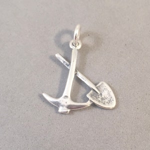 May include: A silver-colored charm featuring a crossed pickaxe and shovel design. The charm has a small loop at the top for attaching to a necklace or bracelet. The tools are detailed with a slightly textured finish.