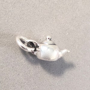 May include: A small, silver-colored teapot charm with a curved handle, spout, and a lid. The charm is detailed and appears to be made of metal. The teapot is set against a gray background.